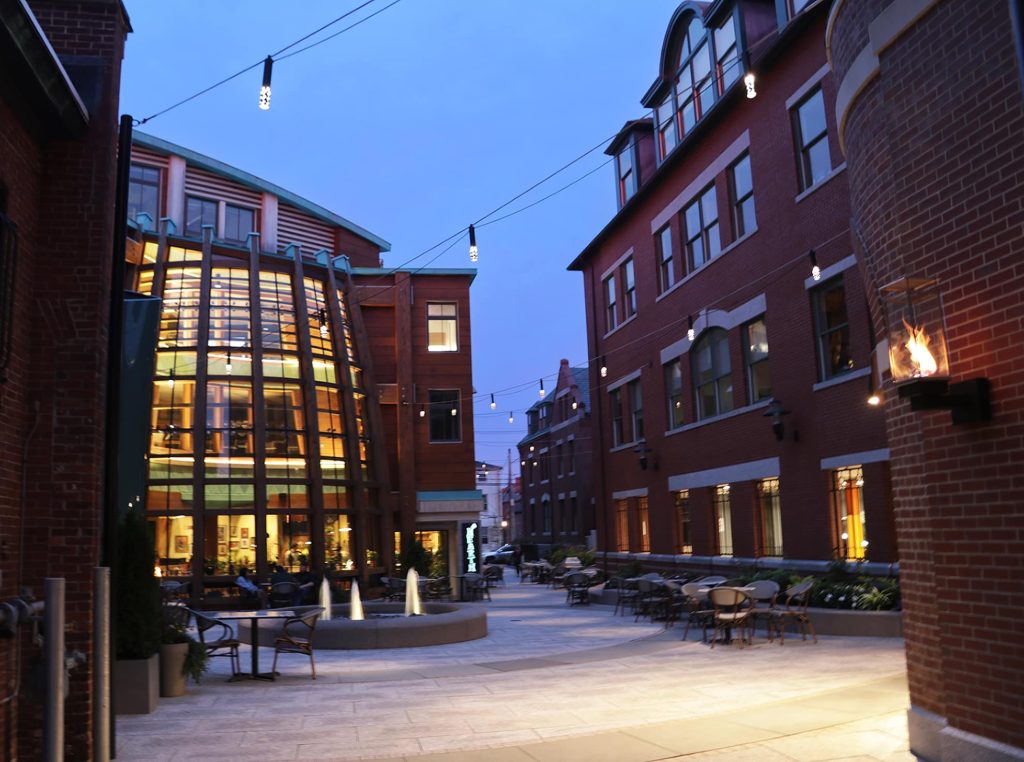 Downtown street at dusk with brick paving, string lights overhead and lit storefronts
