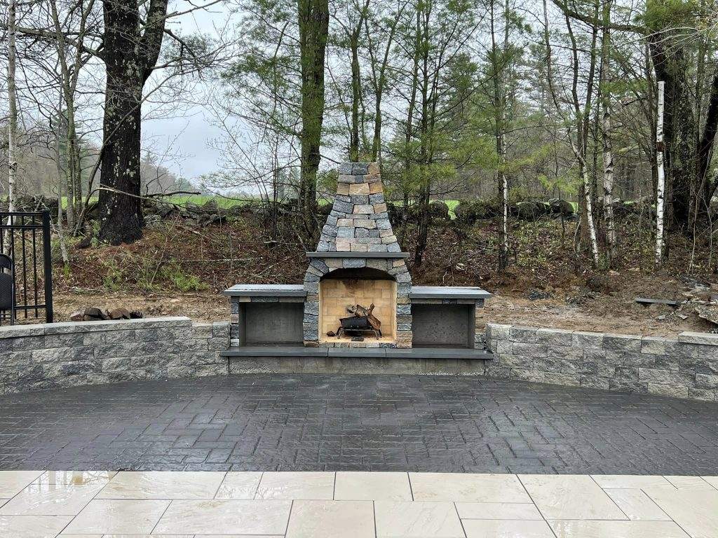 Stone outdoor fireplace built at the edge of a paver patio in a wooded yard